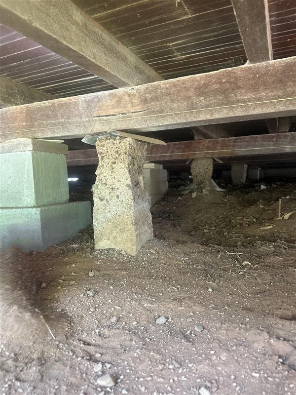 Under raised wooden structure, a crumbling concrete pier supports beams over dirt ground.