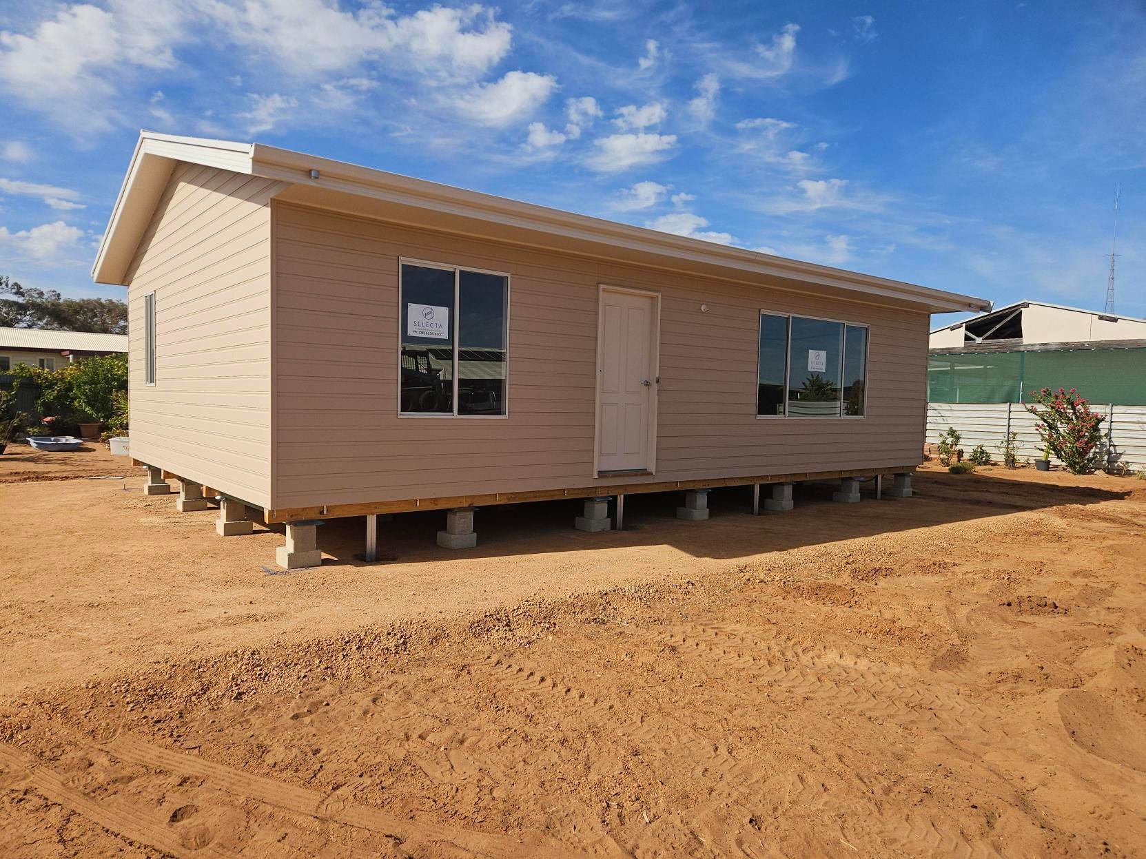 Small beige house under construction on a dirt lot, with windows and a slanted roof under a blue sky. Small beige house under construction on a dirt lot, with windows and a slanted roof under a blue sky.