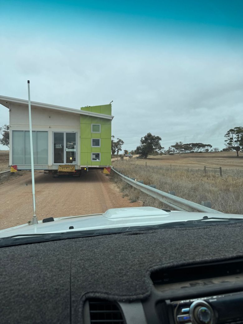 Small green tiny house being towed on a dirt road in a rural field, seen from inside a car.