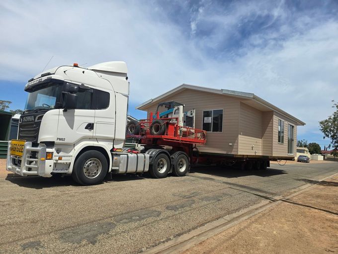 White semi-truck hauling a beige house on a flatbed trailer along a roadside under a blue sky