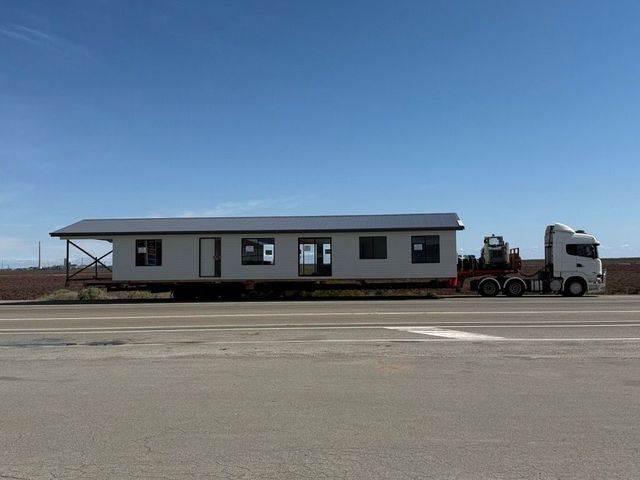 Flatbed truck carrying a gray modular home on a roadside under a clear blue sky.