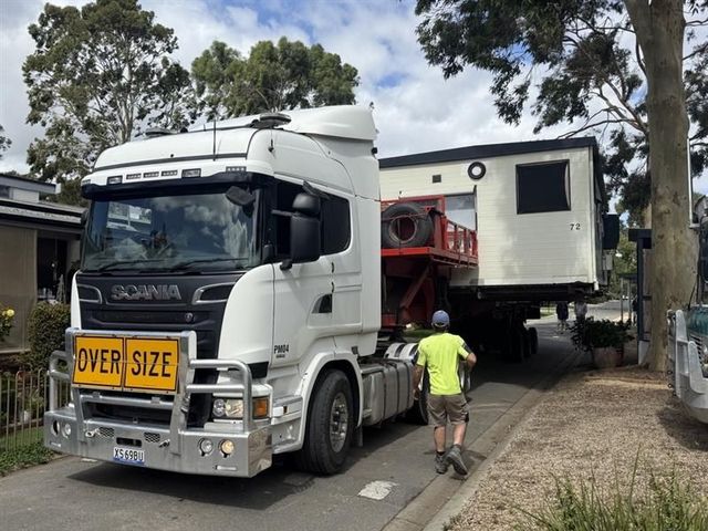 White truck transporting a tiny house, with a worker guiding it and an “OVERSIZE” sign on the front.