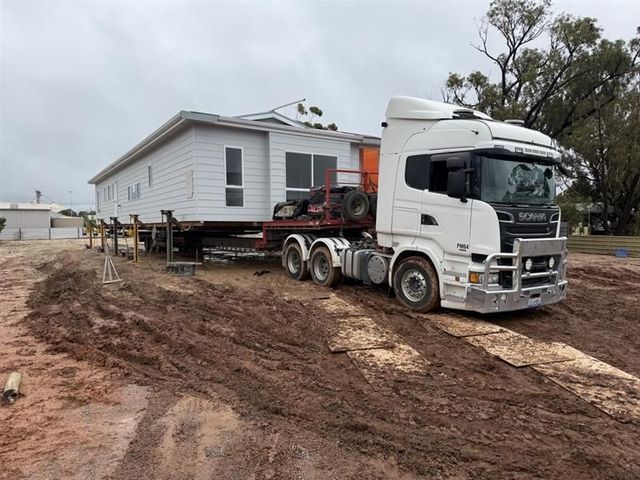 White semi-truck hauling a raised mobile home across a muddy construction site