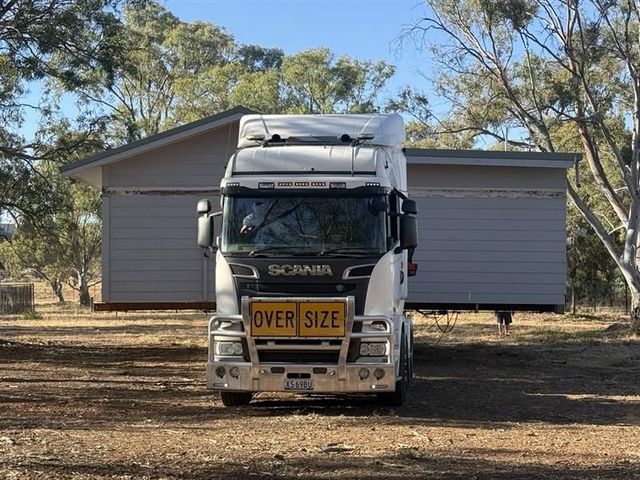 White Scania semi-truck with “OVERSIZE” sign parked in front of a shed in a rural setting