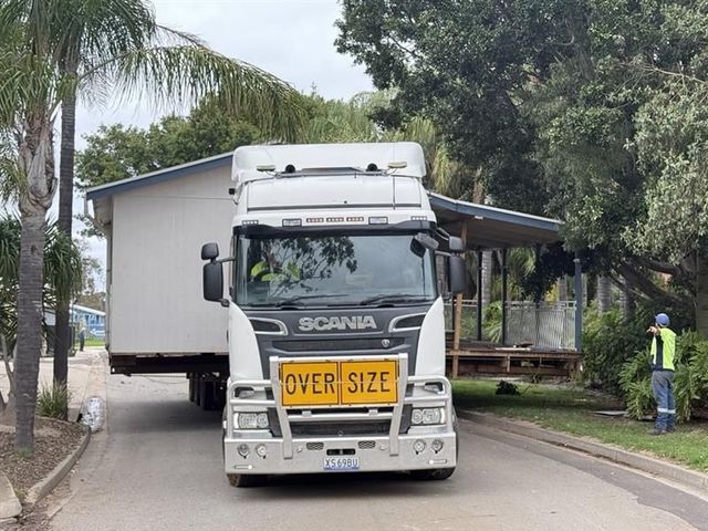 White Scania oversize truck on a road with a yellow “OVER SIZE” sign, trees and a person at right.