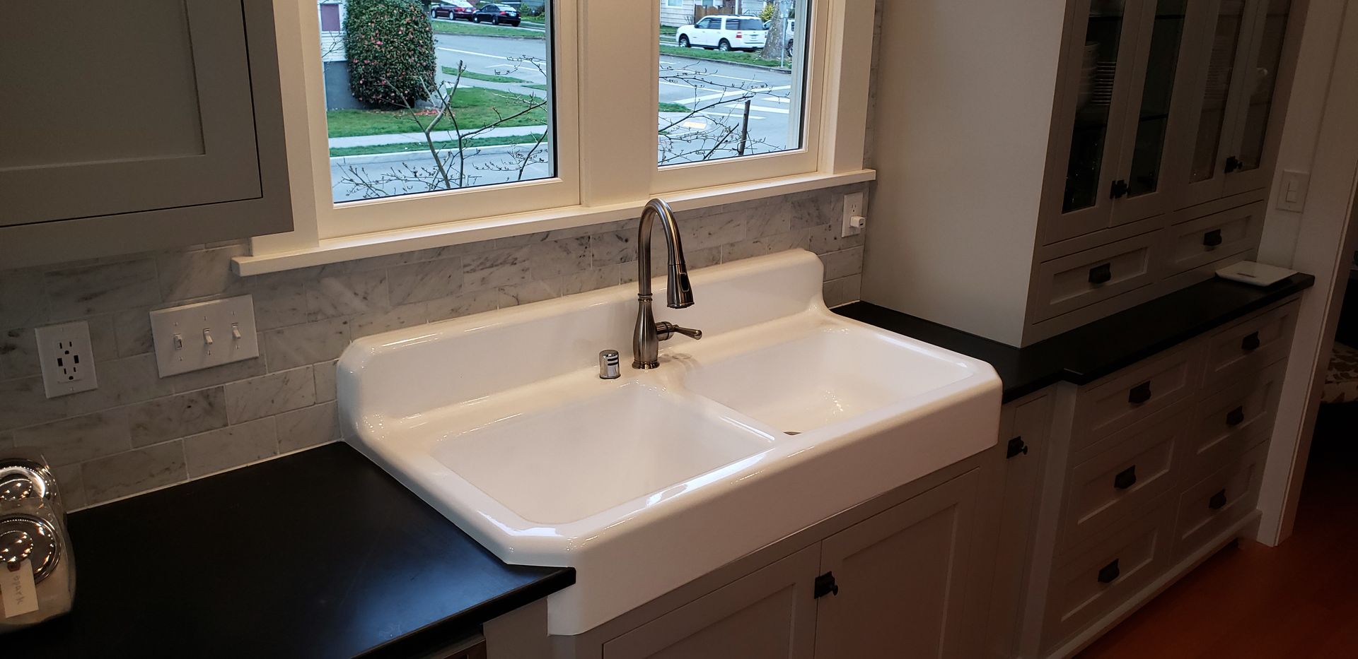 White farmhouse sink in a kitchen, black countertops, cabinets, window, and faucet.
