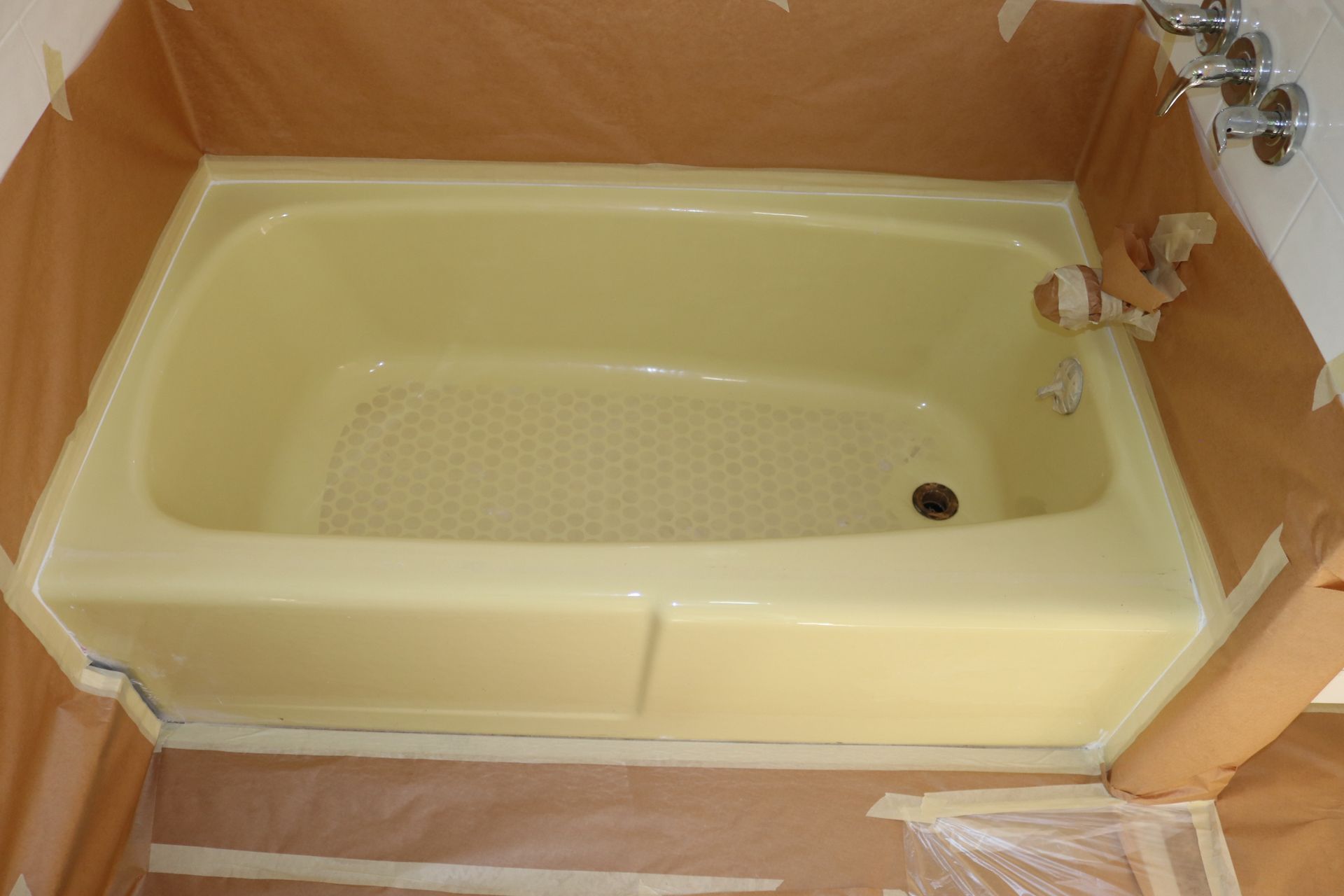 Tan bathtub prepared for refinishing, surrounded by protective paper and tape.