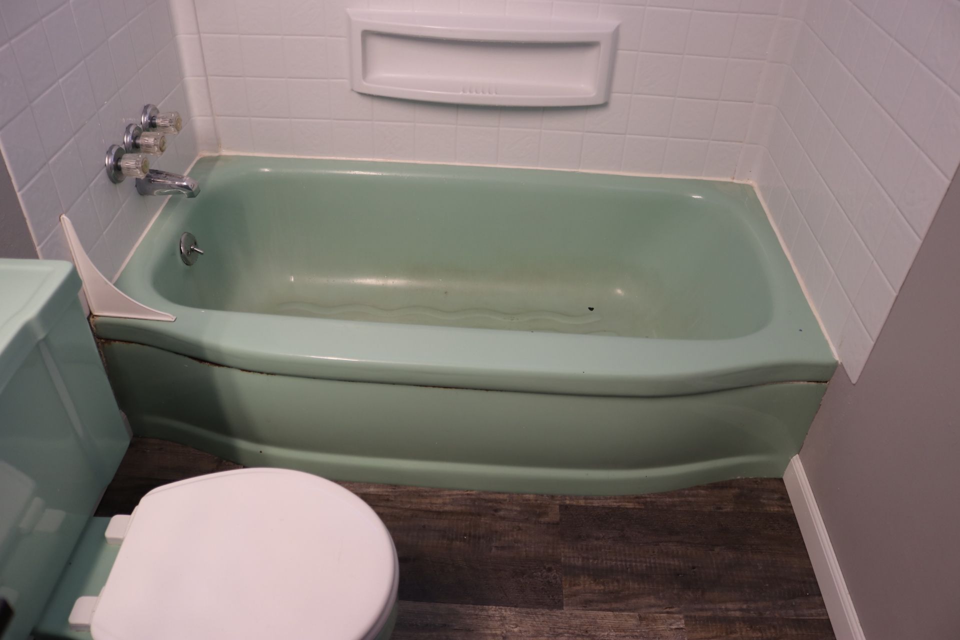 Green bathtub and toilet in bathroom with wood-look flooring and white tile walls.