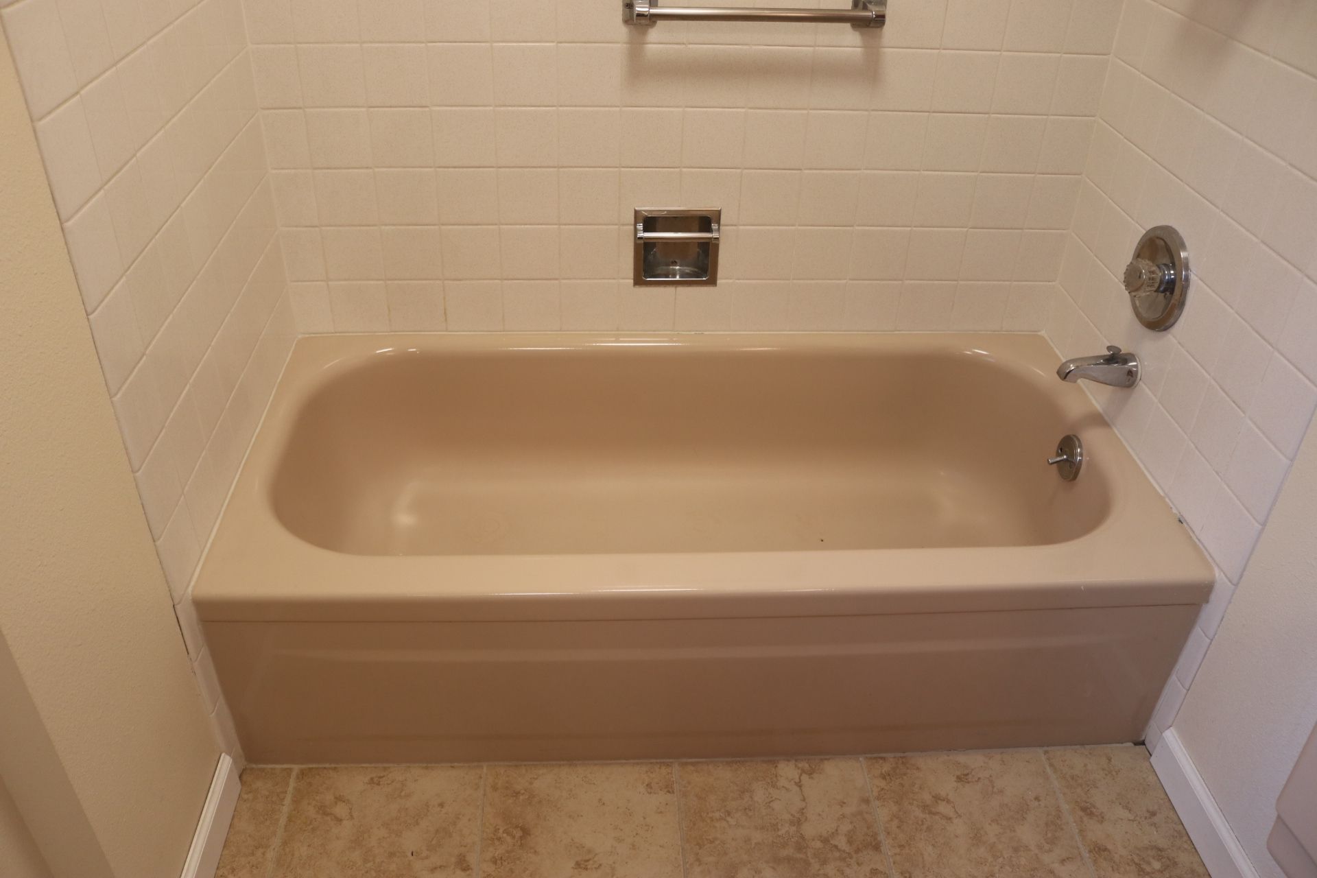 A beige bathtub in a bathroom, surrounded by cream-colored tiled walls and a towel rack.