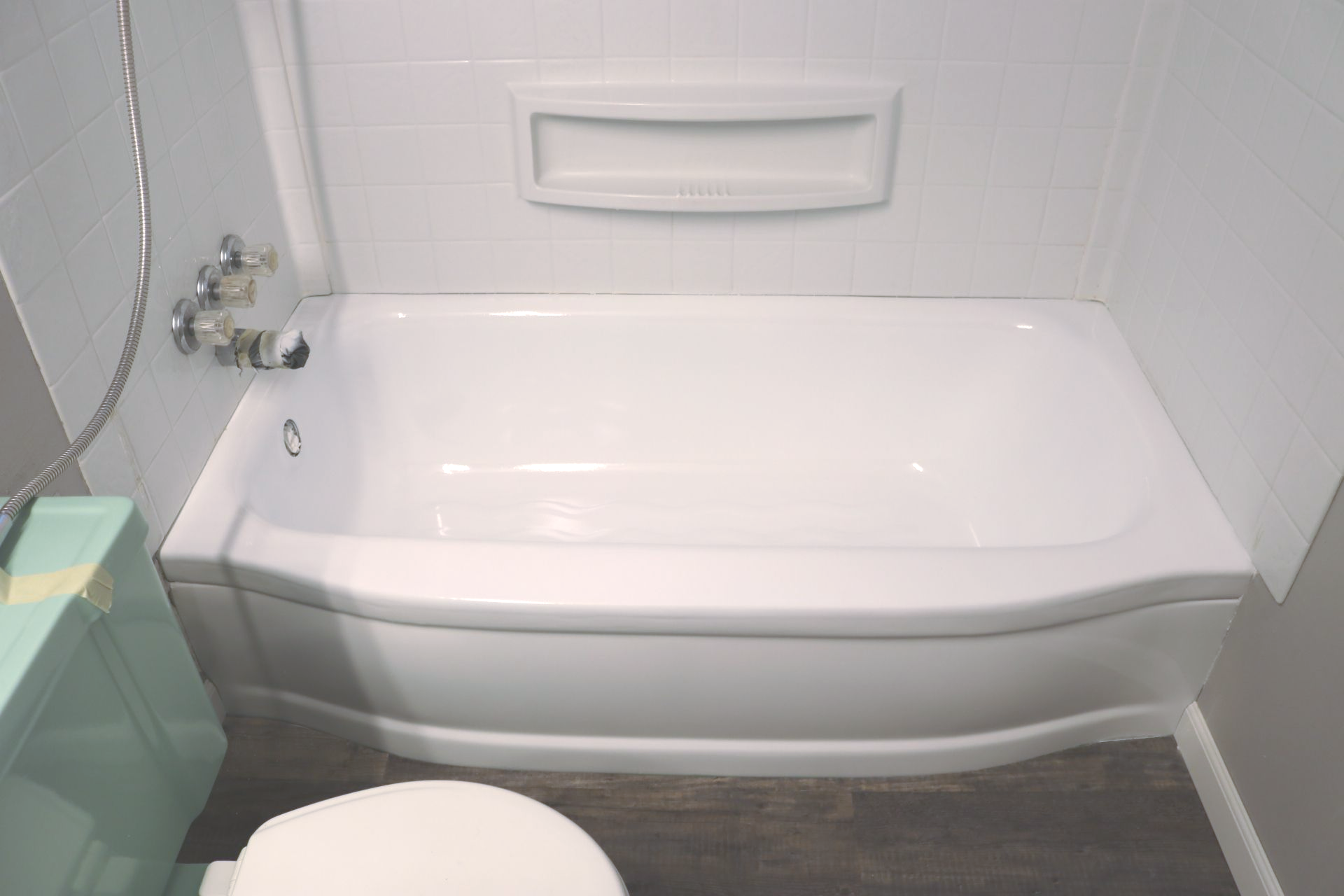 White bathtub with a built-in soap dish, next to a pale blue toilet in a white-tiled bathroom.