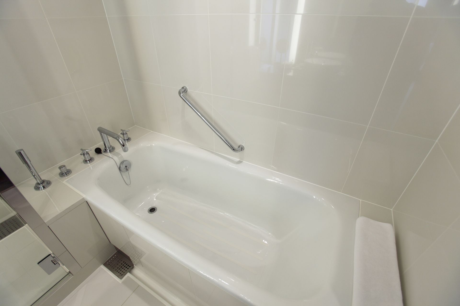 White bathtub with chrome fixtures and grab bar in a tiled bathroom. White bathtub with chrome fixtures and grab bar in a tiled bathroom.