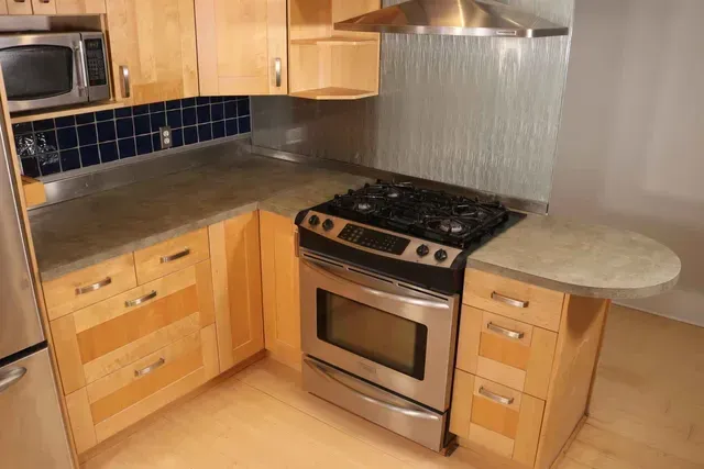 A kitchen with light wood cabinets, stainless steel appliances, and gray countertops.
