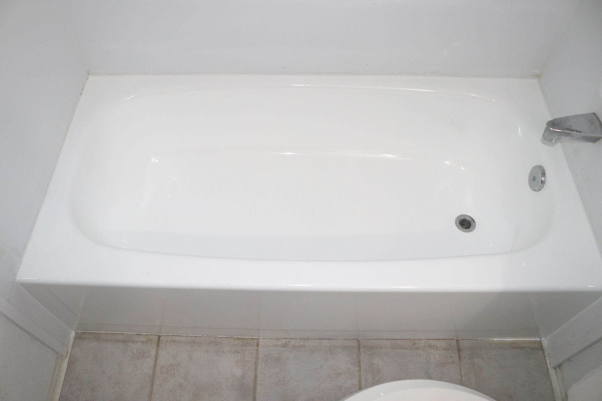 White bathtub built into white-tiled alcove with chrome faucet and drain.
