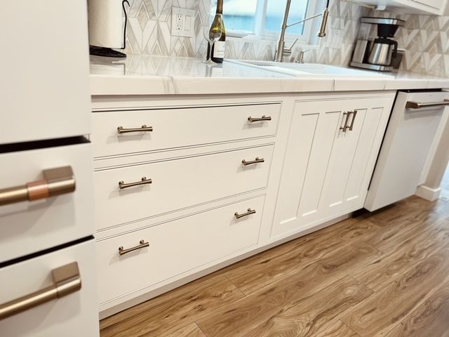 White kitchen cabinets with bronze handles and a white dishwasher. Wooden floor and backsplash.
