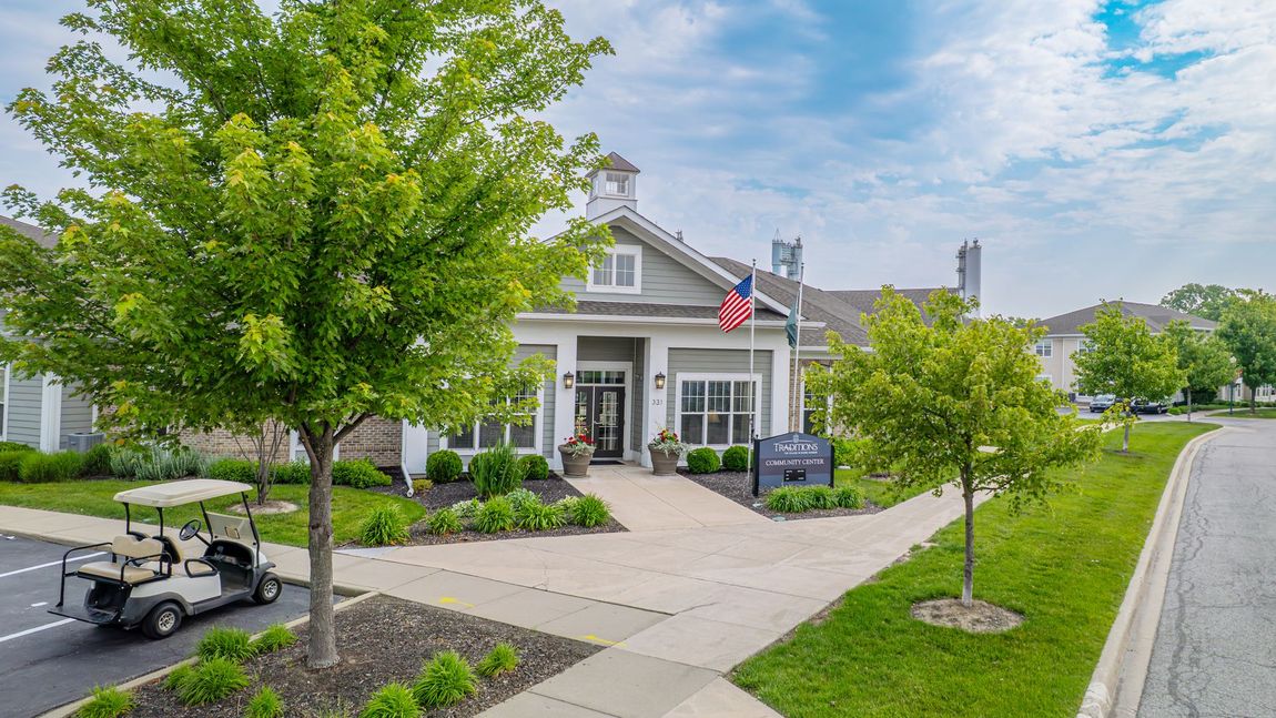 Exterior of a gray building with an American flag. A golf cart sits in the parking lot with trees and green grass.