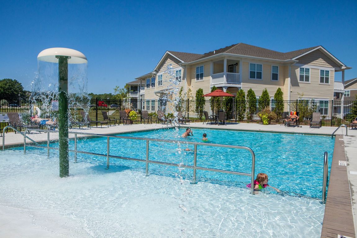 Swimming pool with water fountain and apartment building in the background. Several people in the pool.