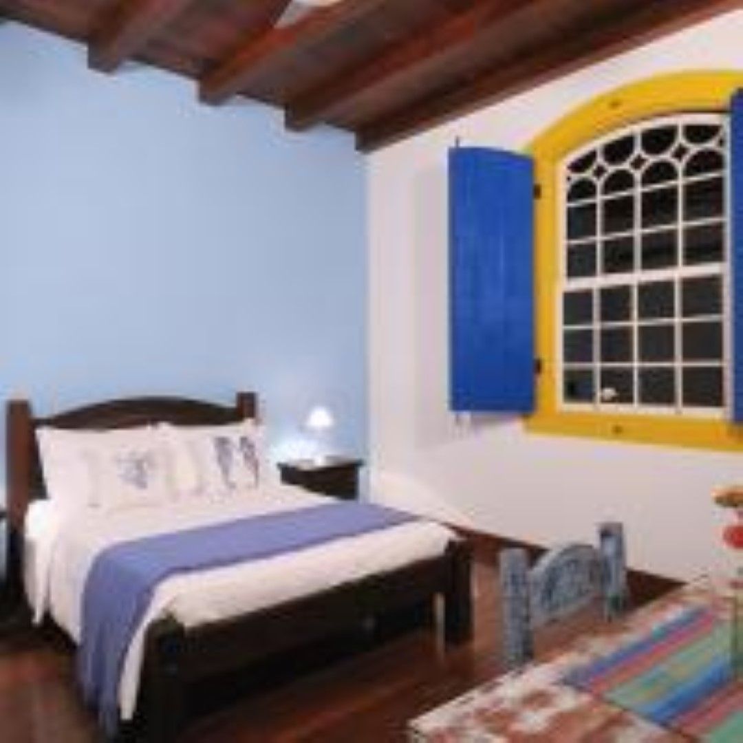 Quarto com parede azul, cama e janela com persianas azuis e moldura amarela. Uma mesa de madeira em primeiro plano.