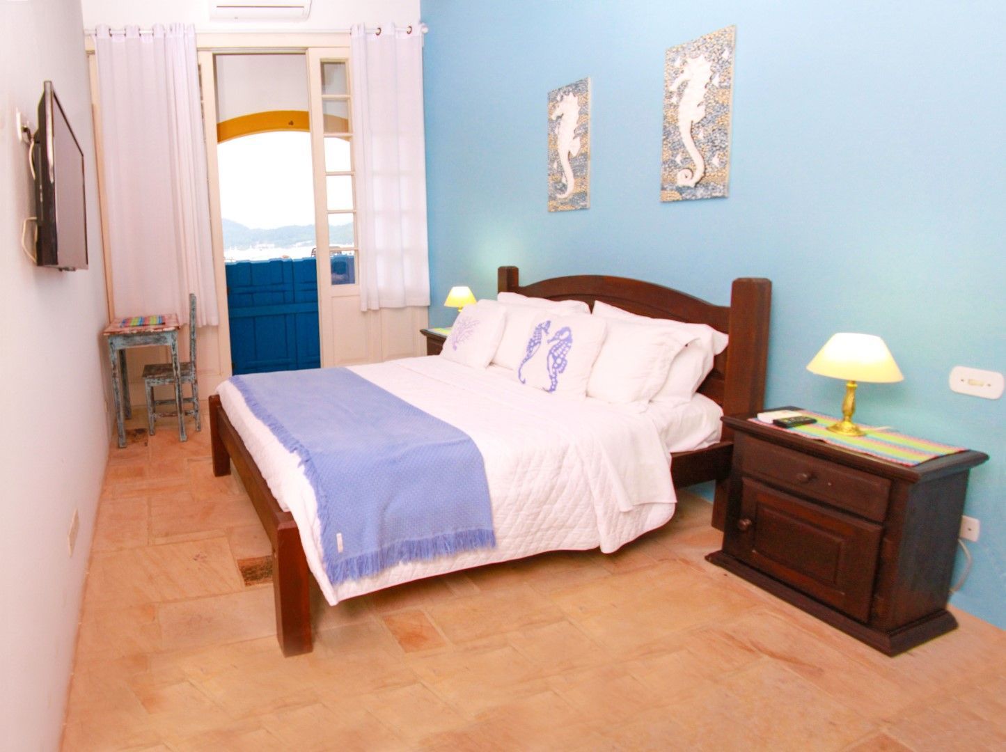 Quarto com parede azul, cama, mesa de cabeceira, mesa pequena, porta para varanda e decoração com cavalos-marinhos.