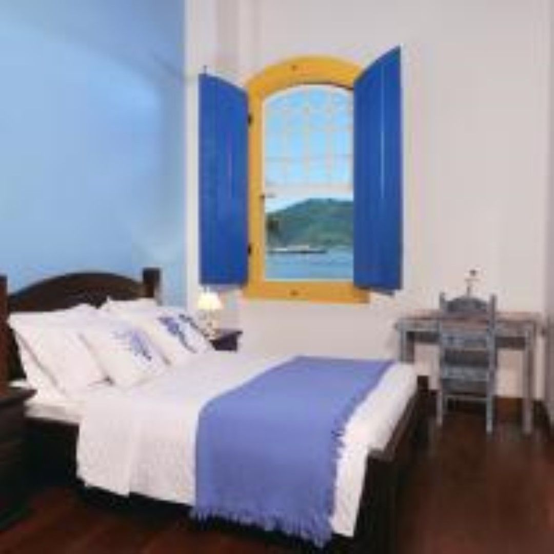Quarto com cama, escrivaninha e uma janela com vista para um corpo d'água; as paredes são azuis e brancas, e as persianas das janelas são azuis.