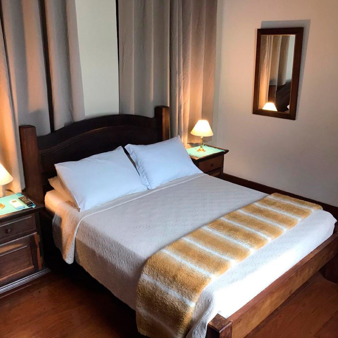 Quarto com cama de madeira, duas mesas de cabeceira com abajures, espelho e roupa de cama bege e branca.
