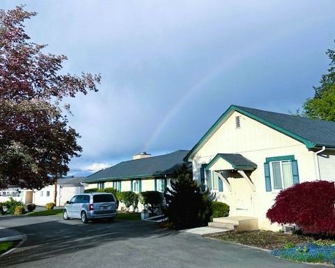 Single-story homes on a quiet street under a rainbow, with a parked silver van and trees.