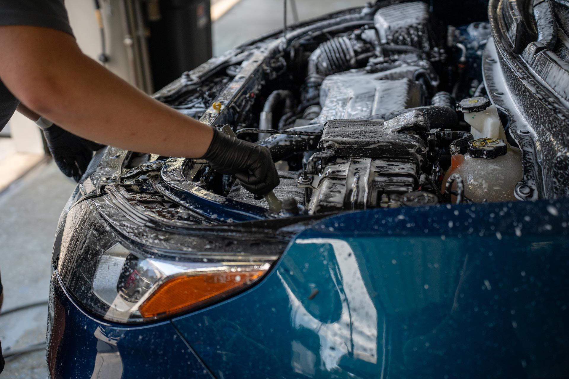 Person cleaning a blue car engine bay with a spray hose at a car wash