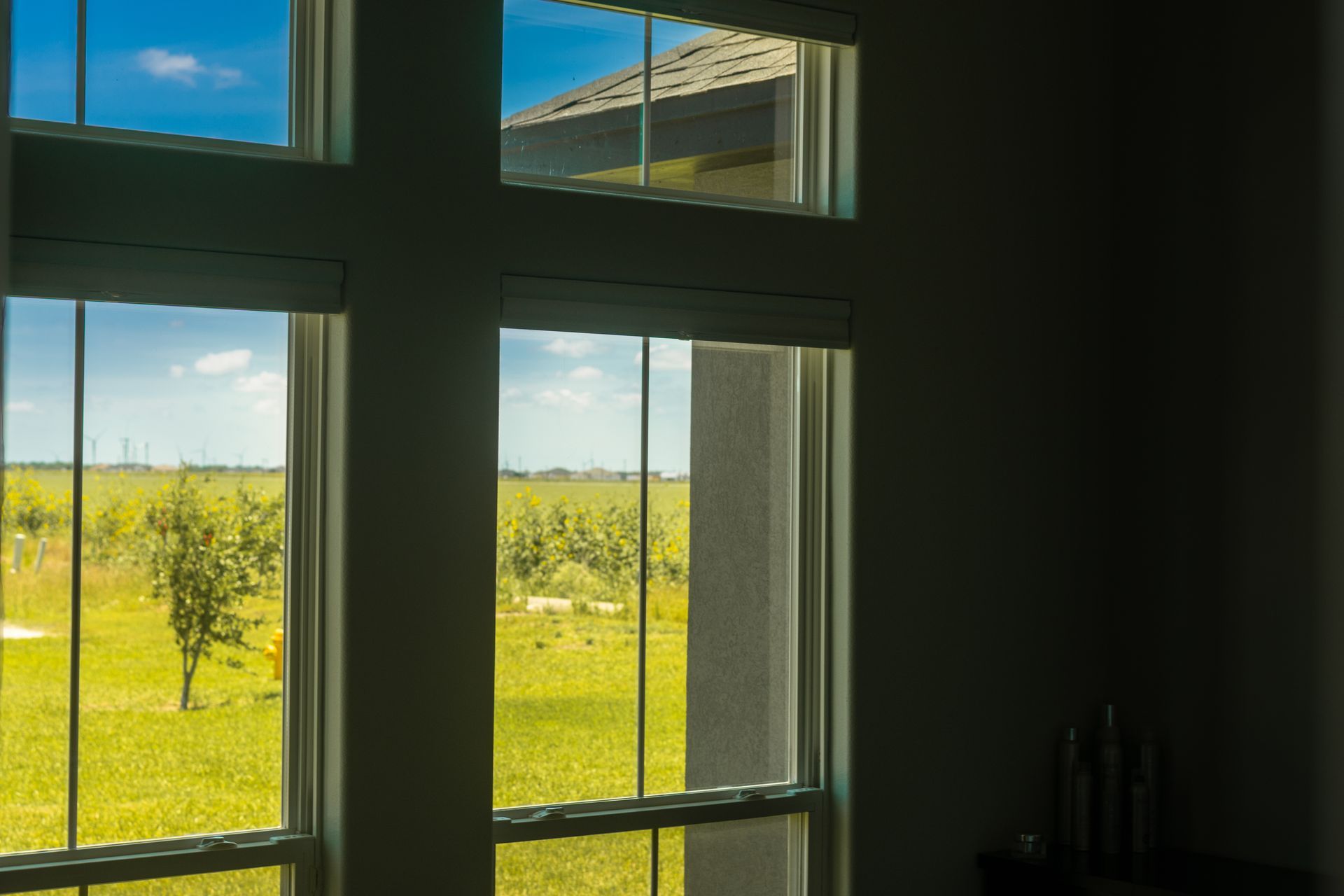 Interior window view of a sunny green field and blue sky, with dark room framing the scene