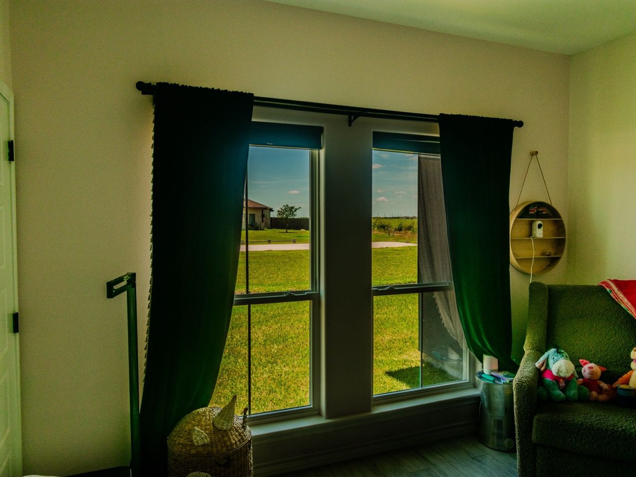 Living room window with black curtains, green lawn outside, and a chair with toys by the wall