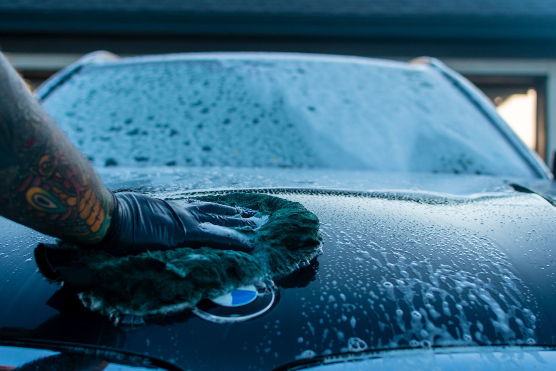 Gloved hand washing a blue car windshield with soap and water droplets