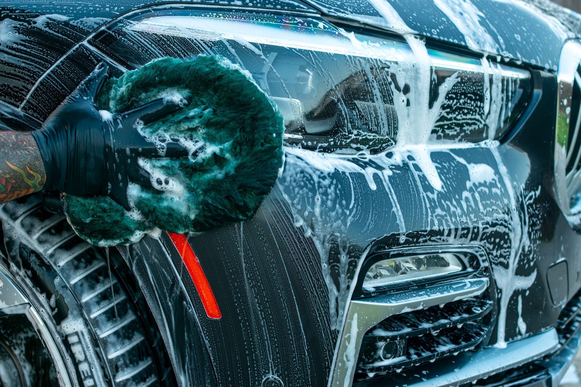 Green microfiber mitt washing a black car grille with soapy foam and water spray