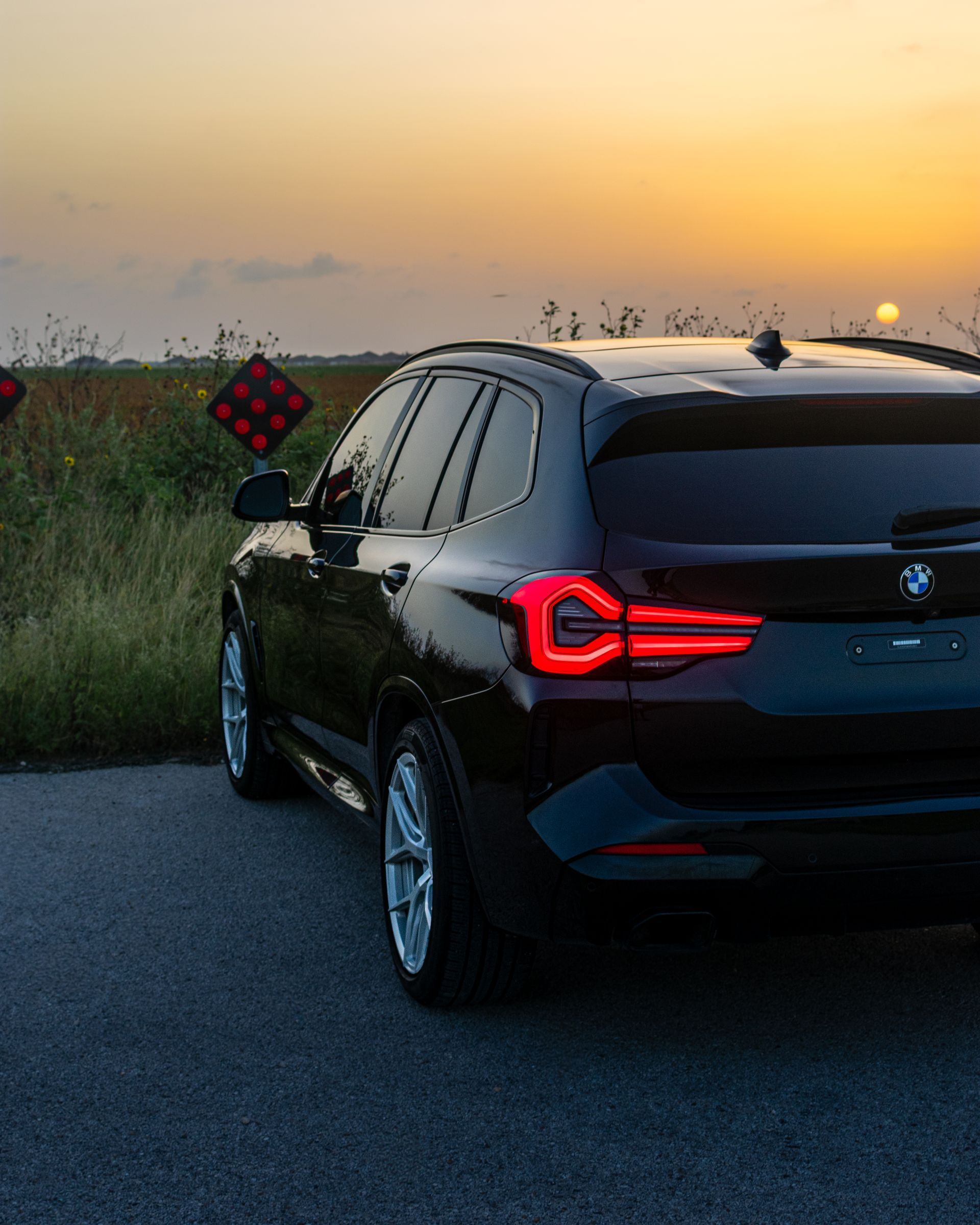 Black SUV parked by roadside at sunset, with glowing red taillights and fields in the background