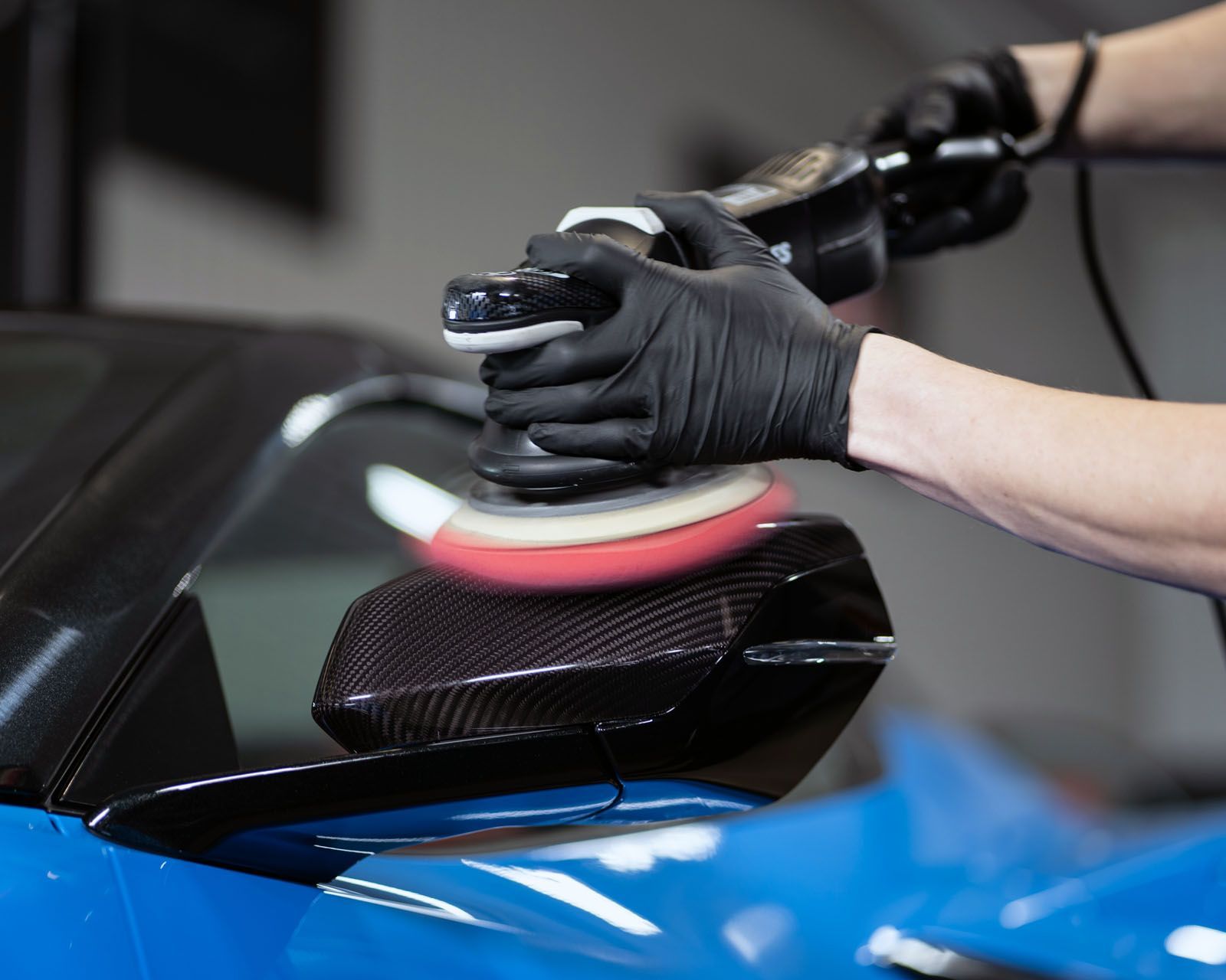 Gloved hands polishing a glossy black car panel with a buffer in an auto detailing shop