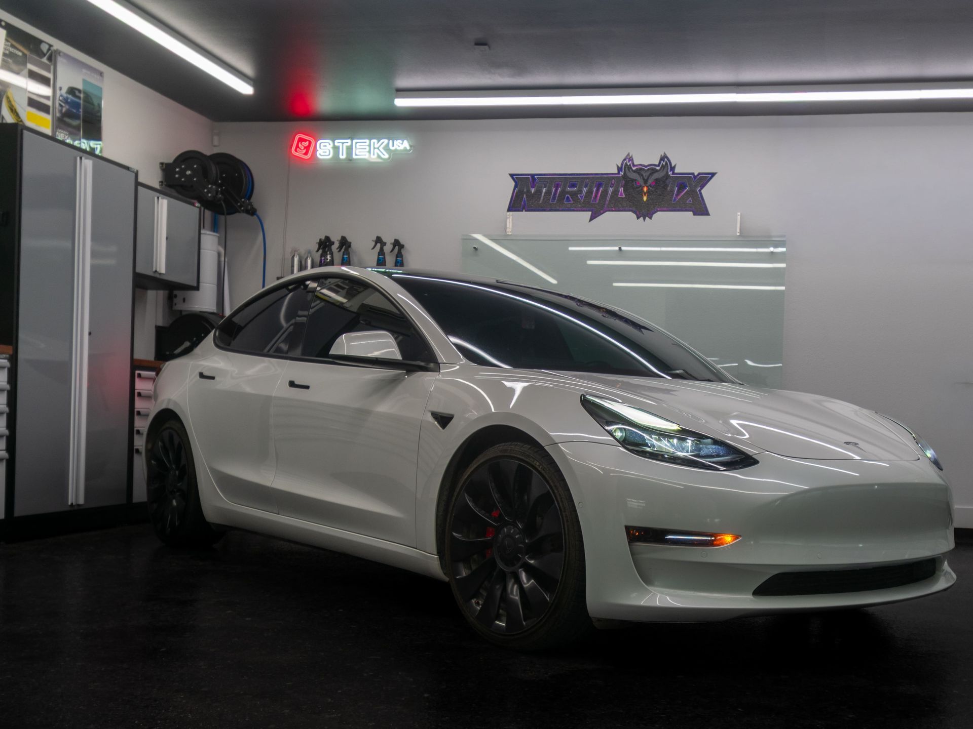 White Tesla sedan in a showroom, parked beside display cabinets under bright ceiling lights.
