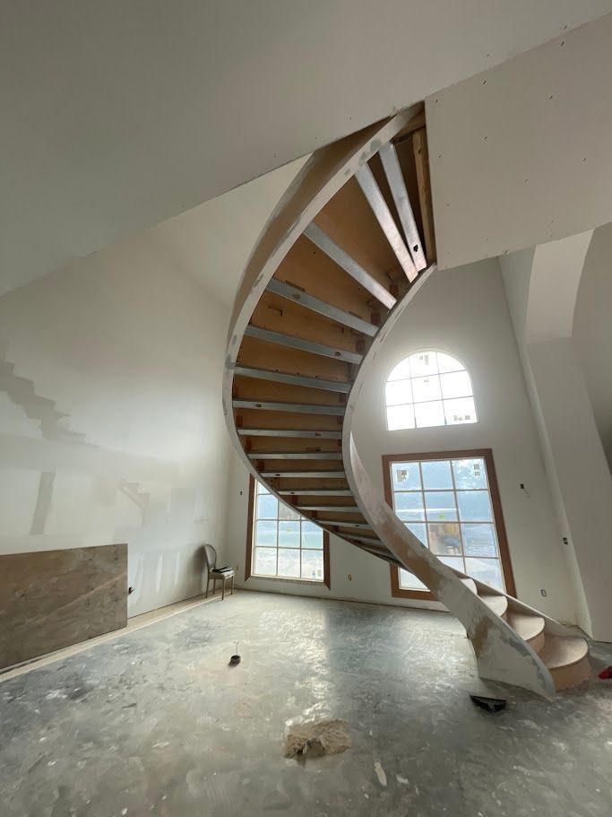 Curved wooden staircase in a bright, unfinished room with large arched windows
