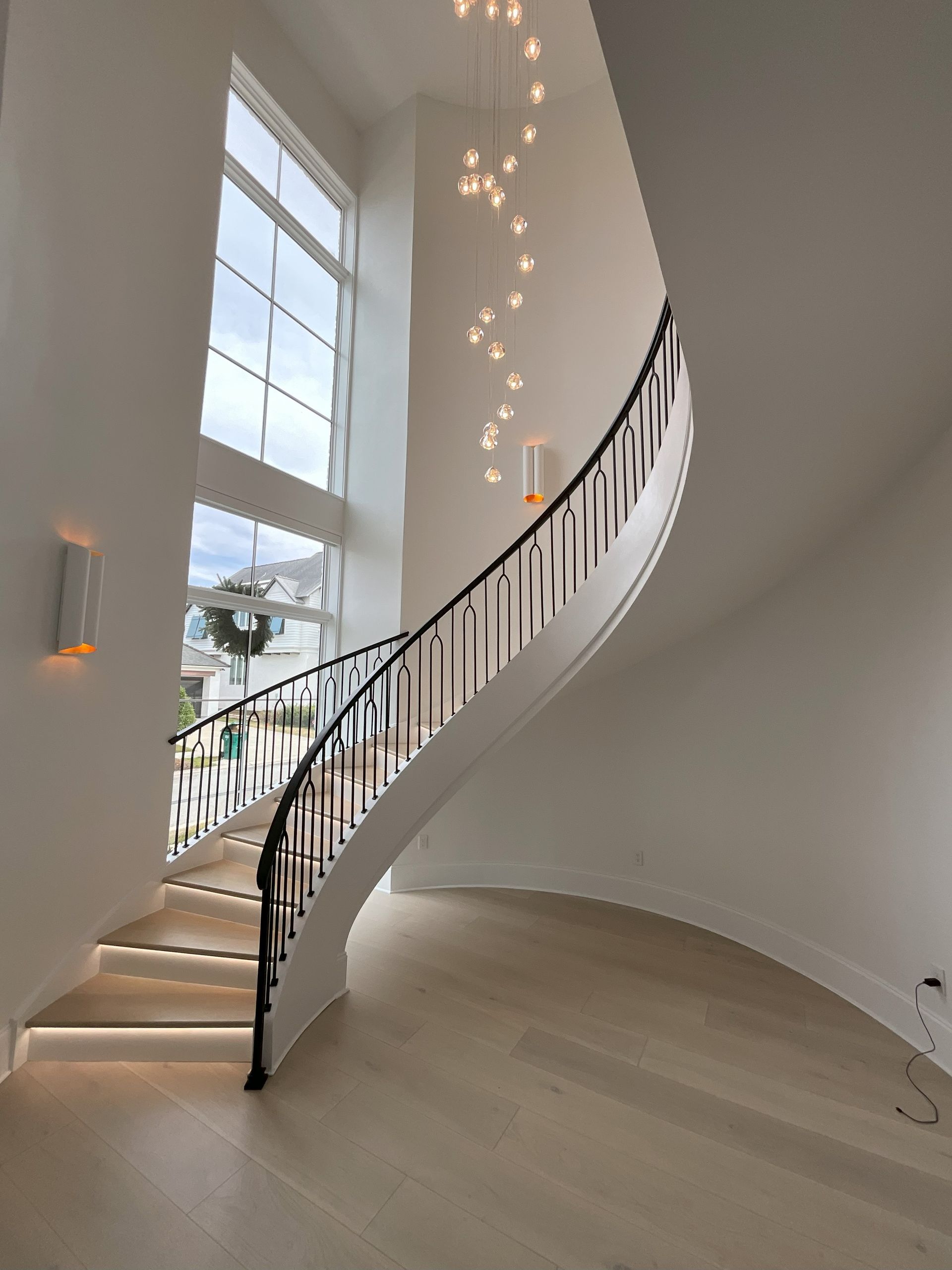 Bright curved staircase with black railing, tall windows, and hanging lights in a white modern interior