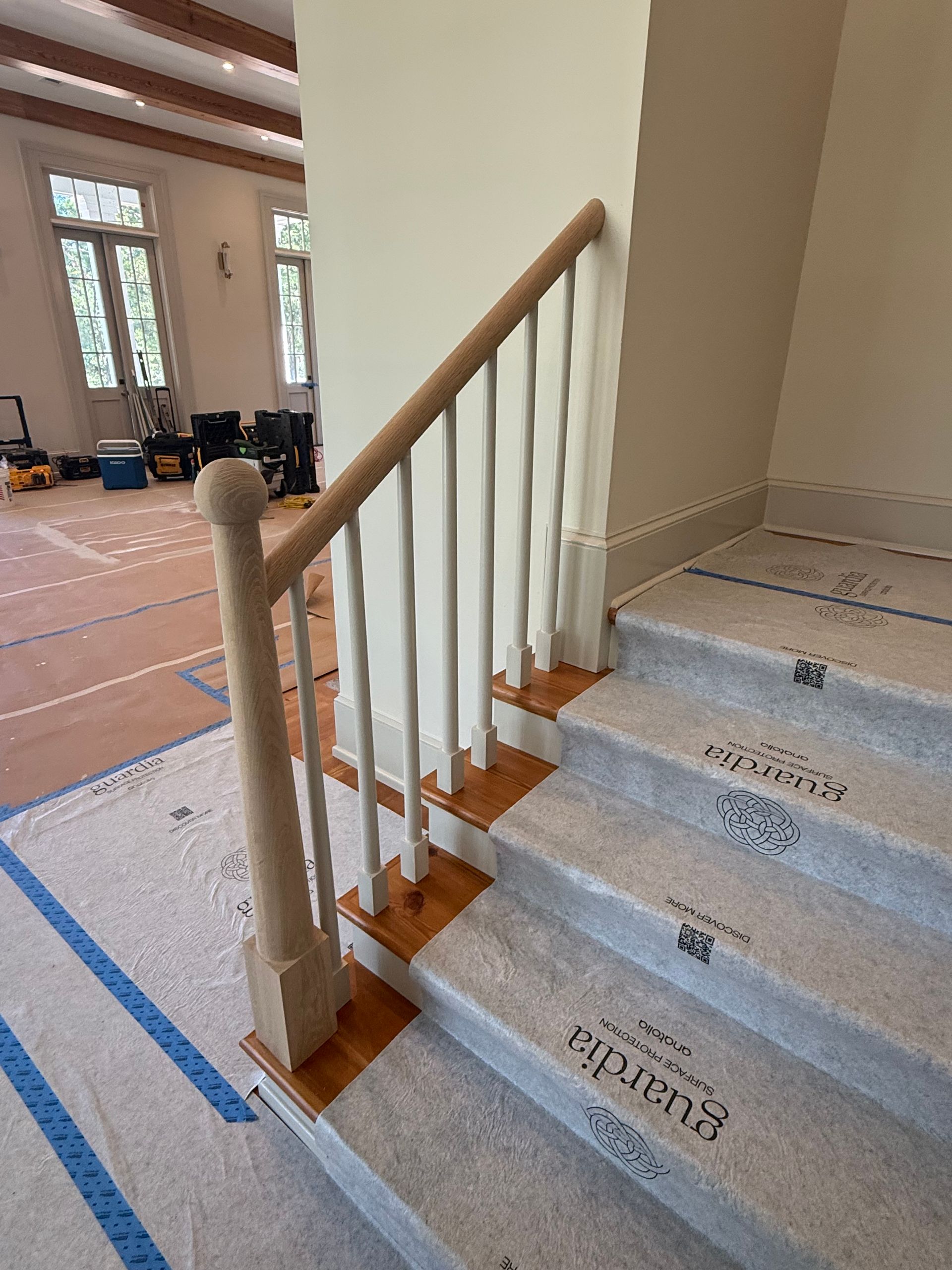 Staircase under renovation with blue tape, plywood floor protection, and a wooden handrail with white balusters.