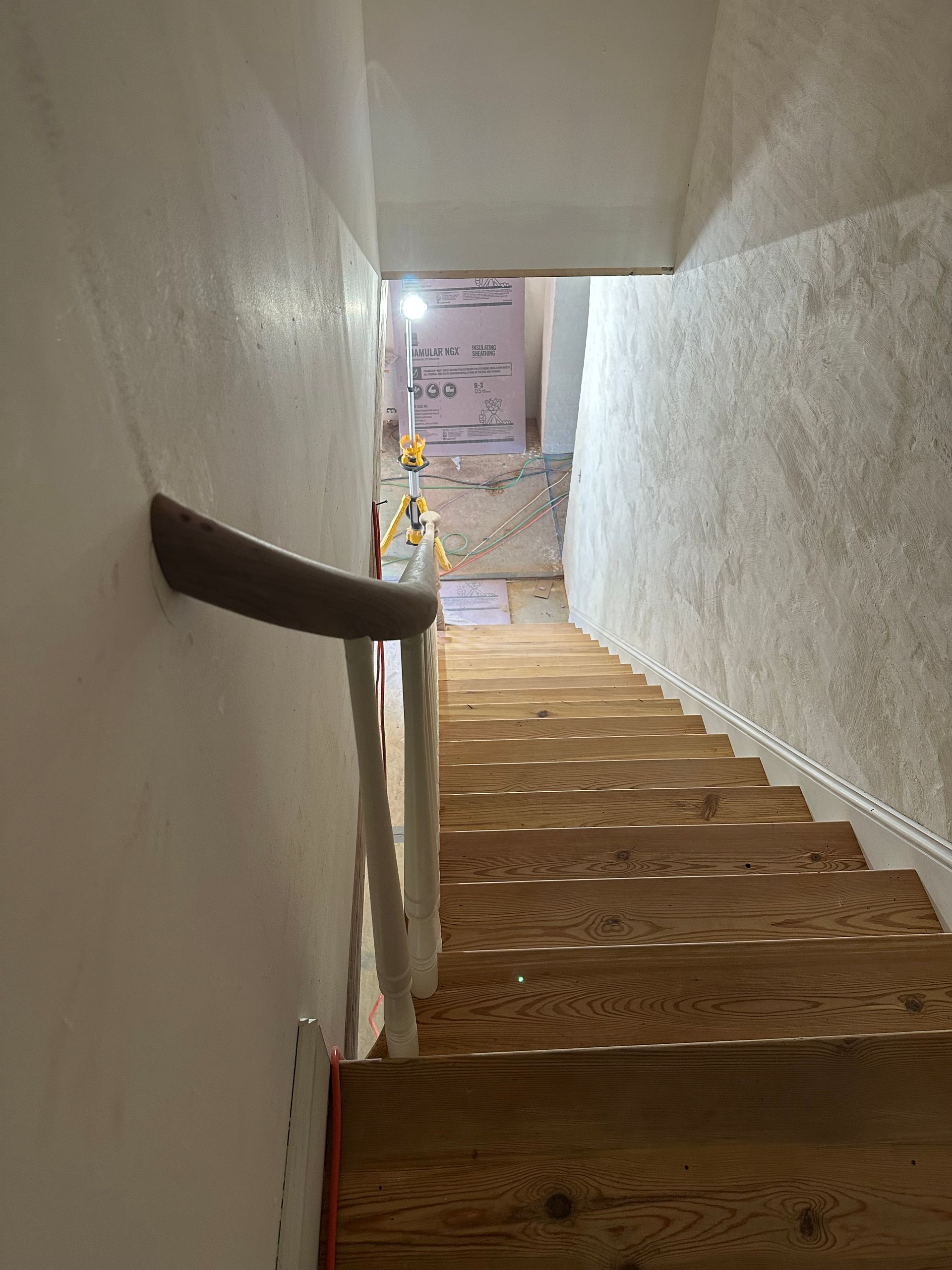 Narrow wooden staircase descending to a bright basement hallway with white walls and a handrail
