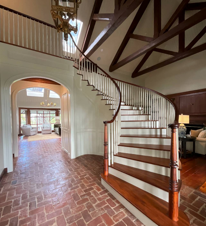 Grand foyer with curved staircase, brick floor, white walls, and exposed dark wood beams