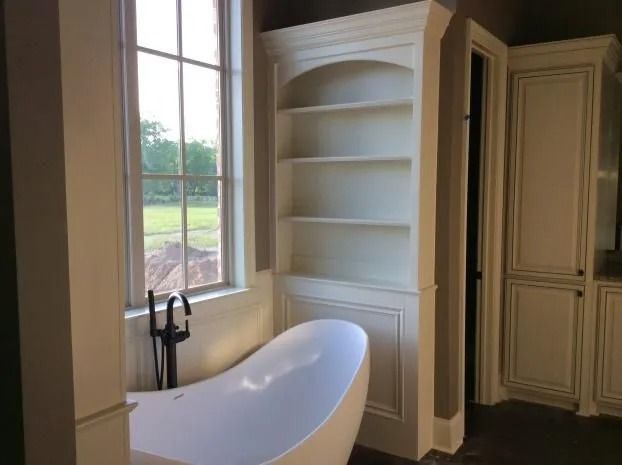 Bathroom with white freestanding tub, built-in shelves, and window overlooking greenery.