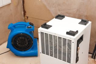 Blue blower and white portable air conditioner on the floor against a cardboard background.