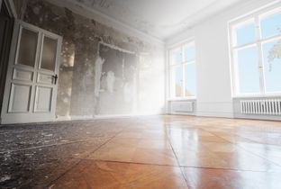 Empty, sunlit room with worn wooden floor, peeling walls, and large windows