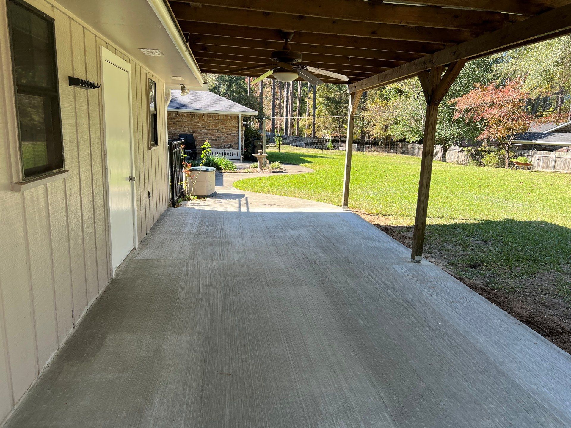 Covered concrete patio beside a house, with lawn and trees in the sunny backyard.