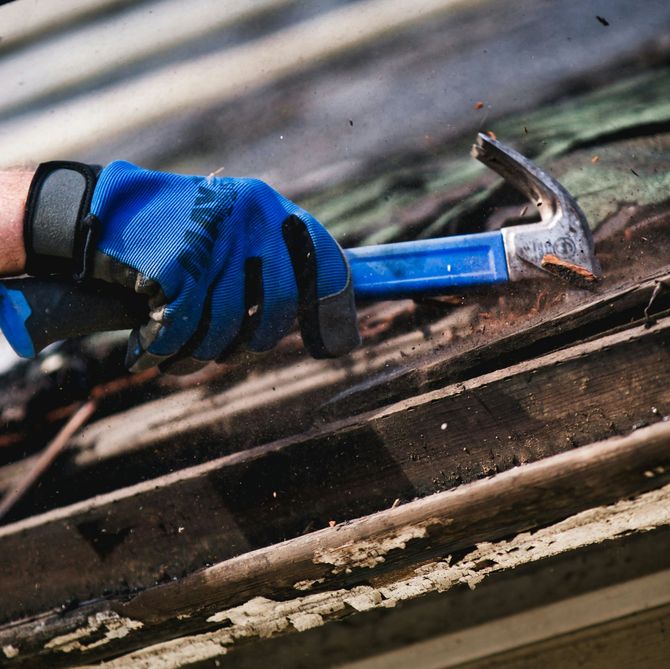 Gloved hand using a blue hammer to remove nails from weathered wood decking.