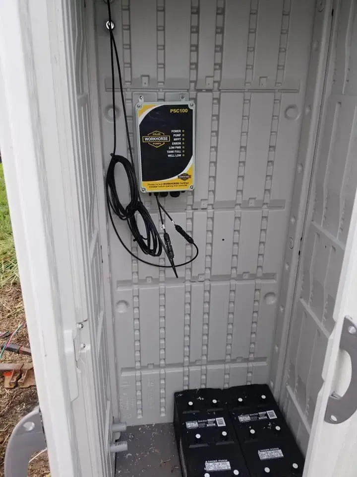 Inside a gray plastic shed: battery charger with cables hanging, two battery boxes on the floor.