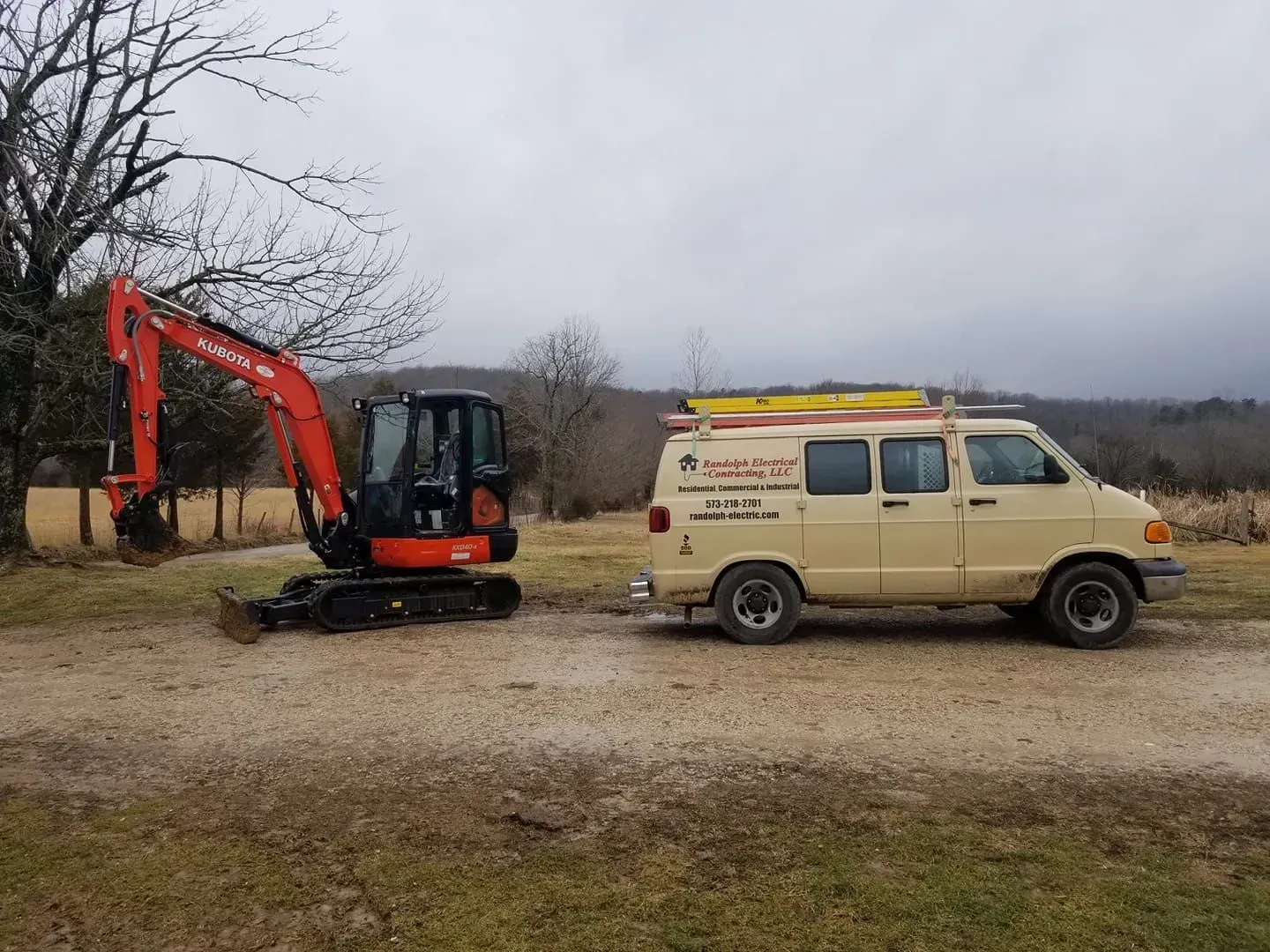 Orange excavator next to a tan work van parked on a gravel road near a field under a cloudy sky.