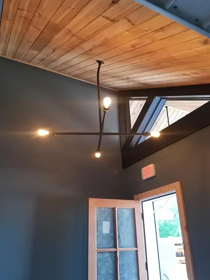 Modern light fixture with four lights against a wood ceiling and dark blue walls, near an open door.