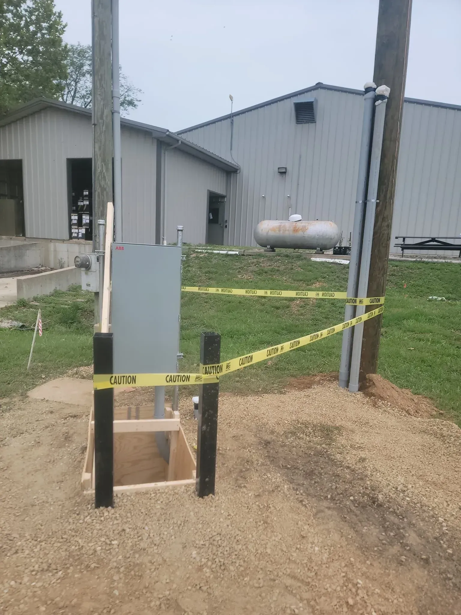 Construction site with gray utility box, poles, caution tape, gravel, and a building in the background.