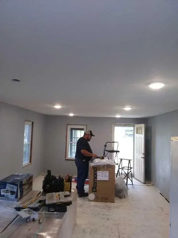Man in a room with tools and boxes. Gray walls, recessed lights. Door to the outside.