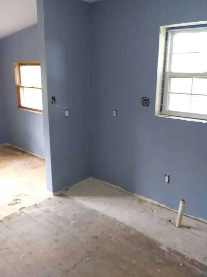 Blue-painted room under construction; two windows, concrete floor, electrical boxes, and pipes.