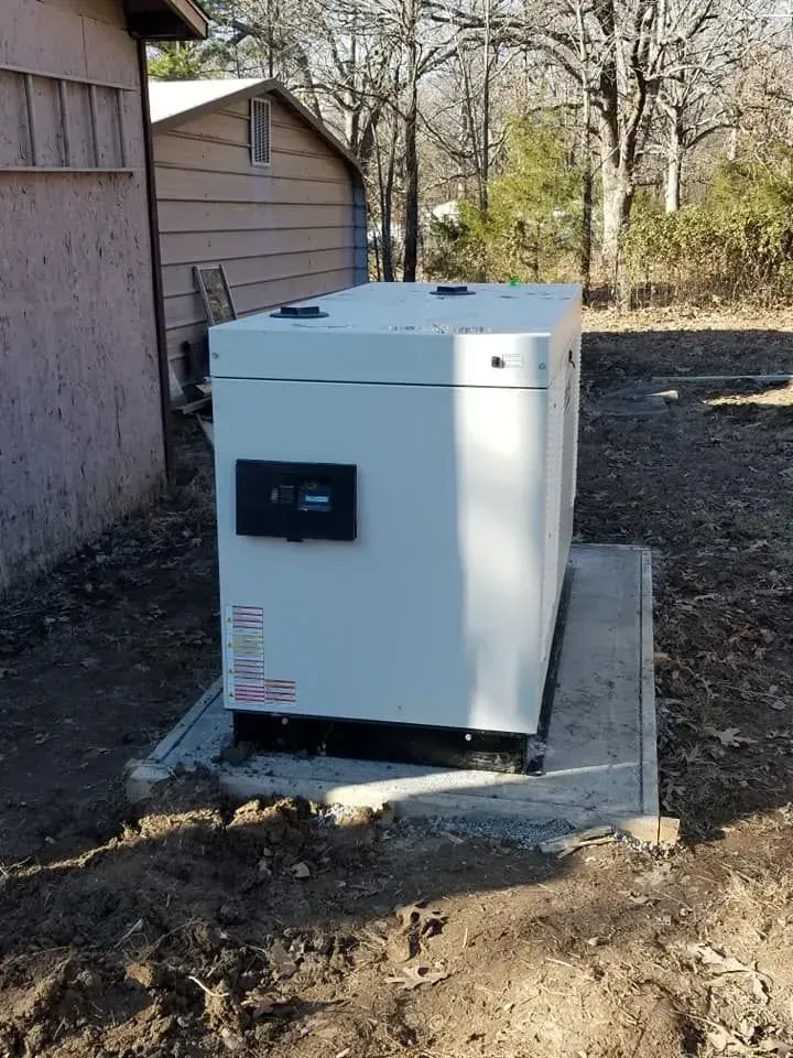 A large, off-white generator on a concrete pad next to a building and shed, outdoors.